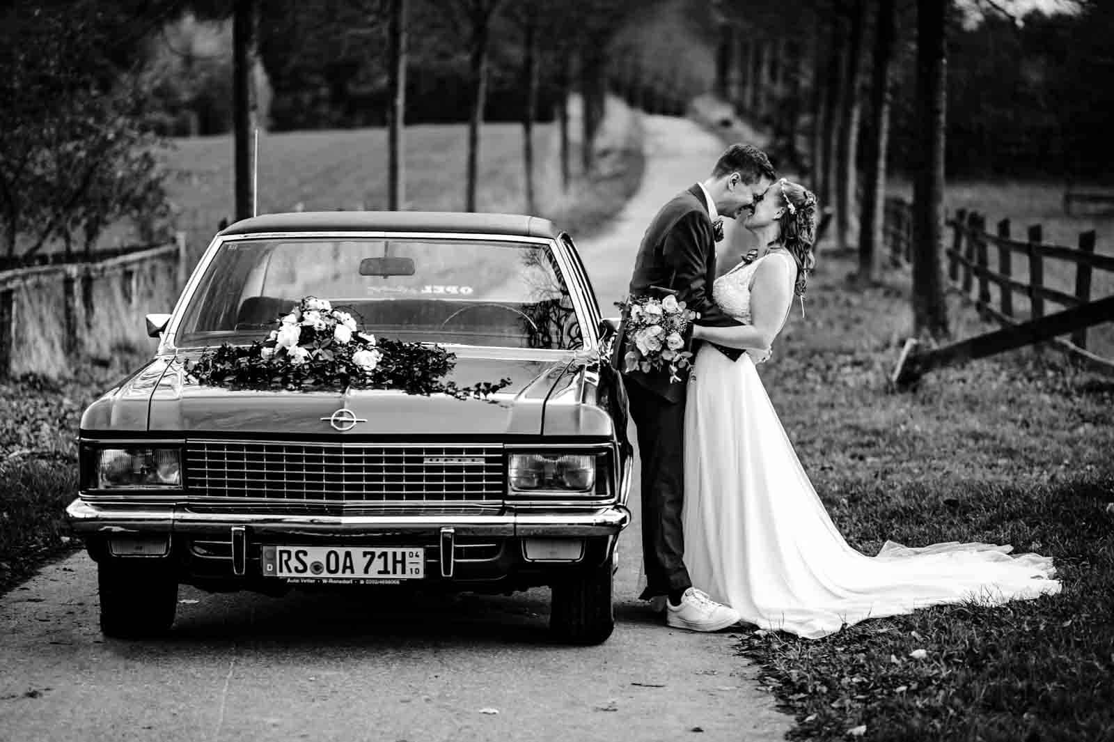 Hochzeitspaar lehnt am Brautauto und küssen sich. Bild in Schwarz Weiß.
